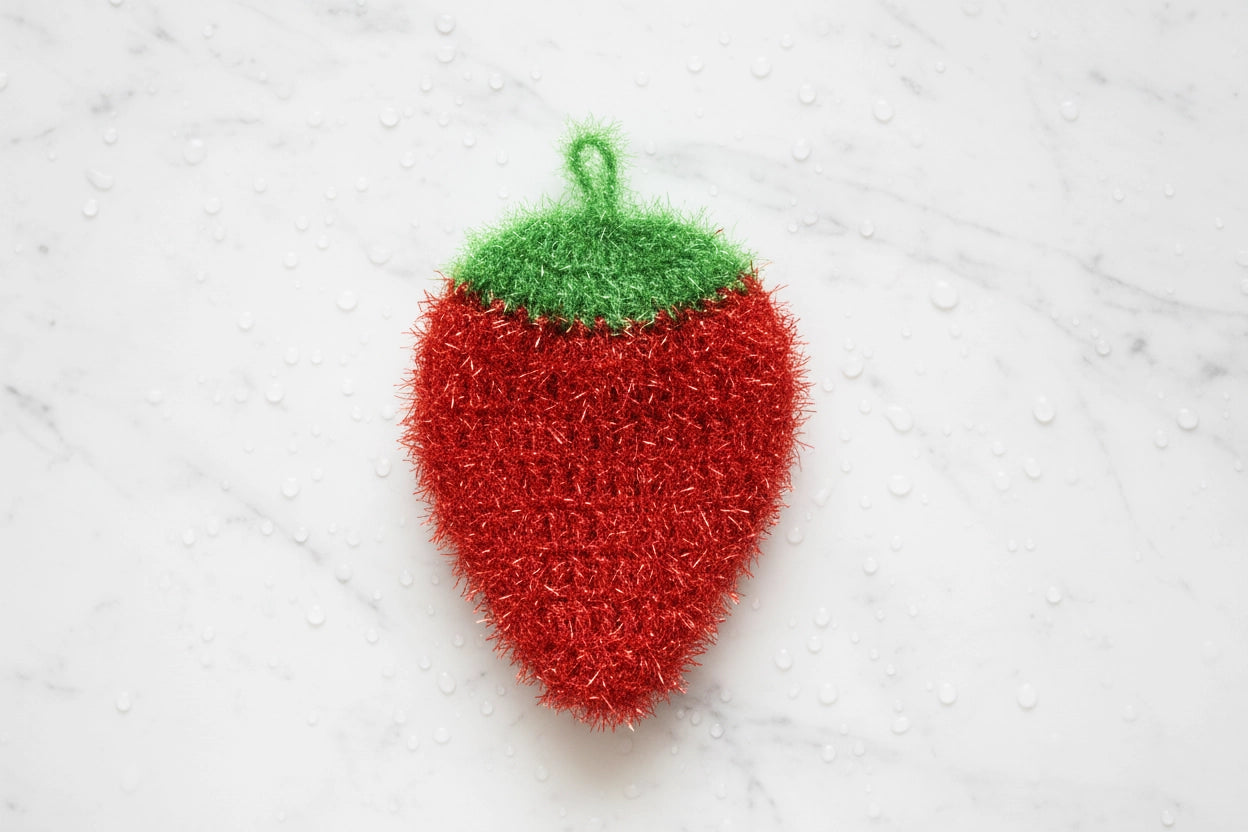Red strawberry-shaped dish scrubber with a green top on a white textured background