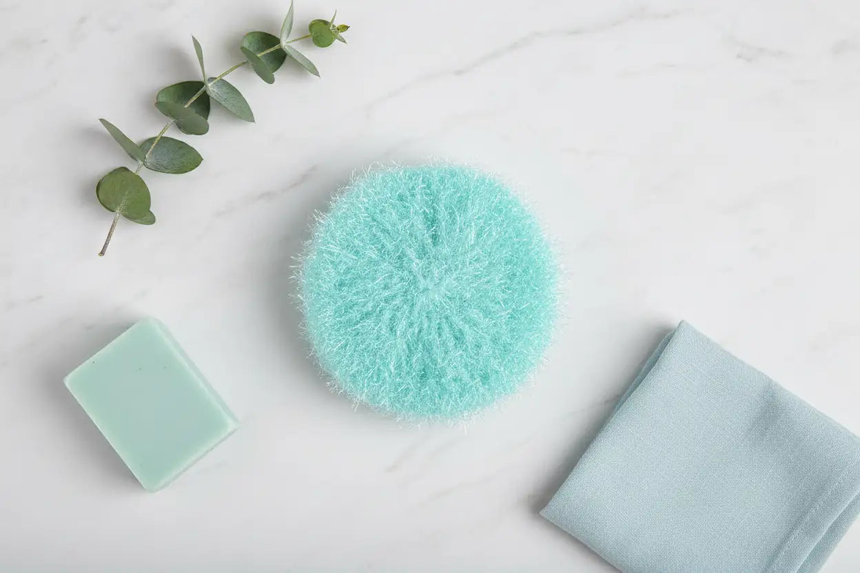 Aqua Hello Scrubby, bar of soap, and towel on a light surface with greenery.