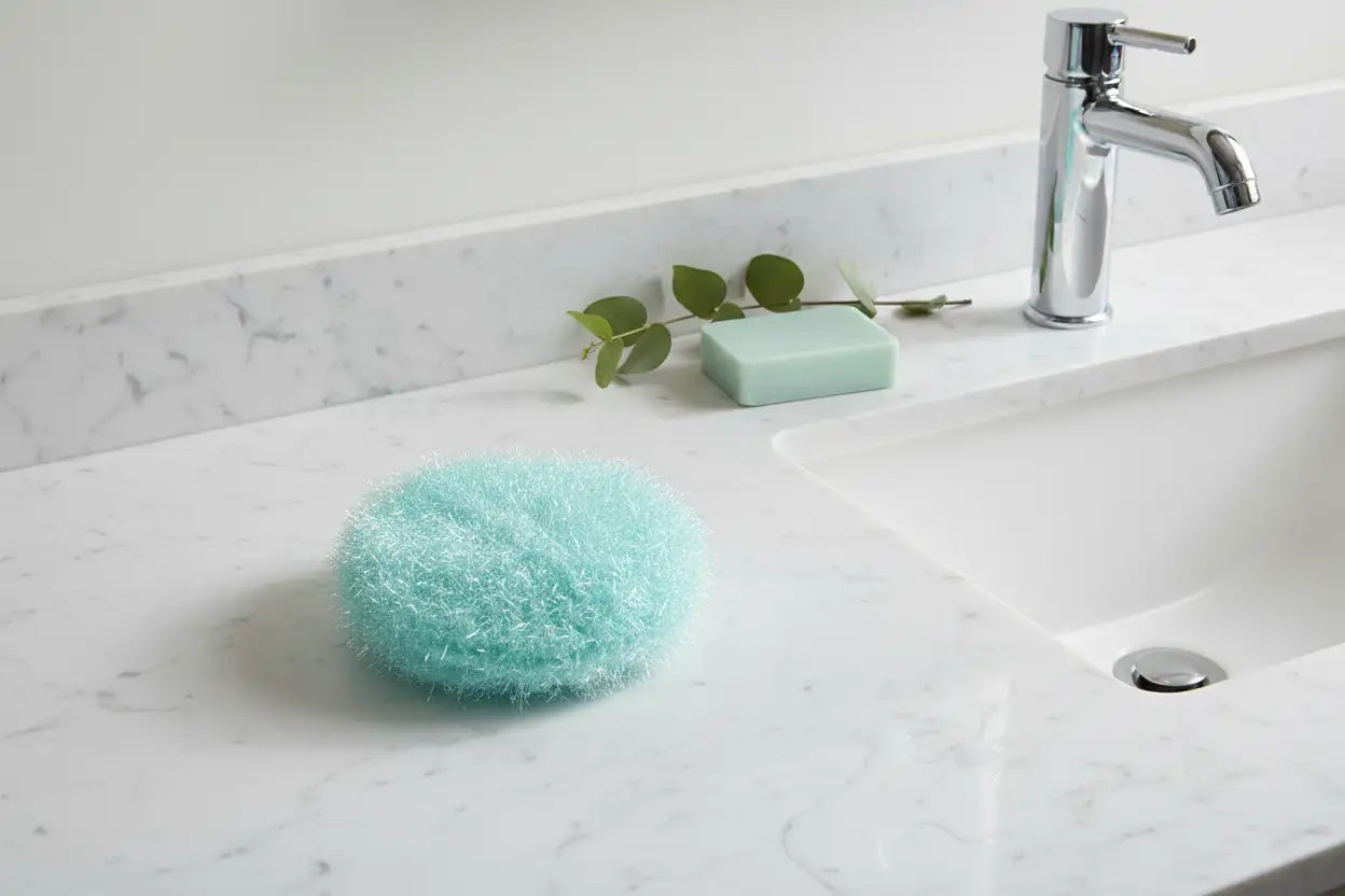 Aqua Hello Scrubby on a marble countertop with a sink and faucet in the background.