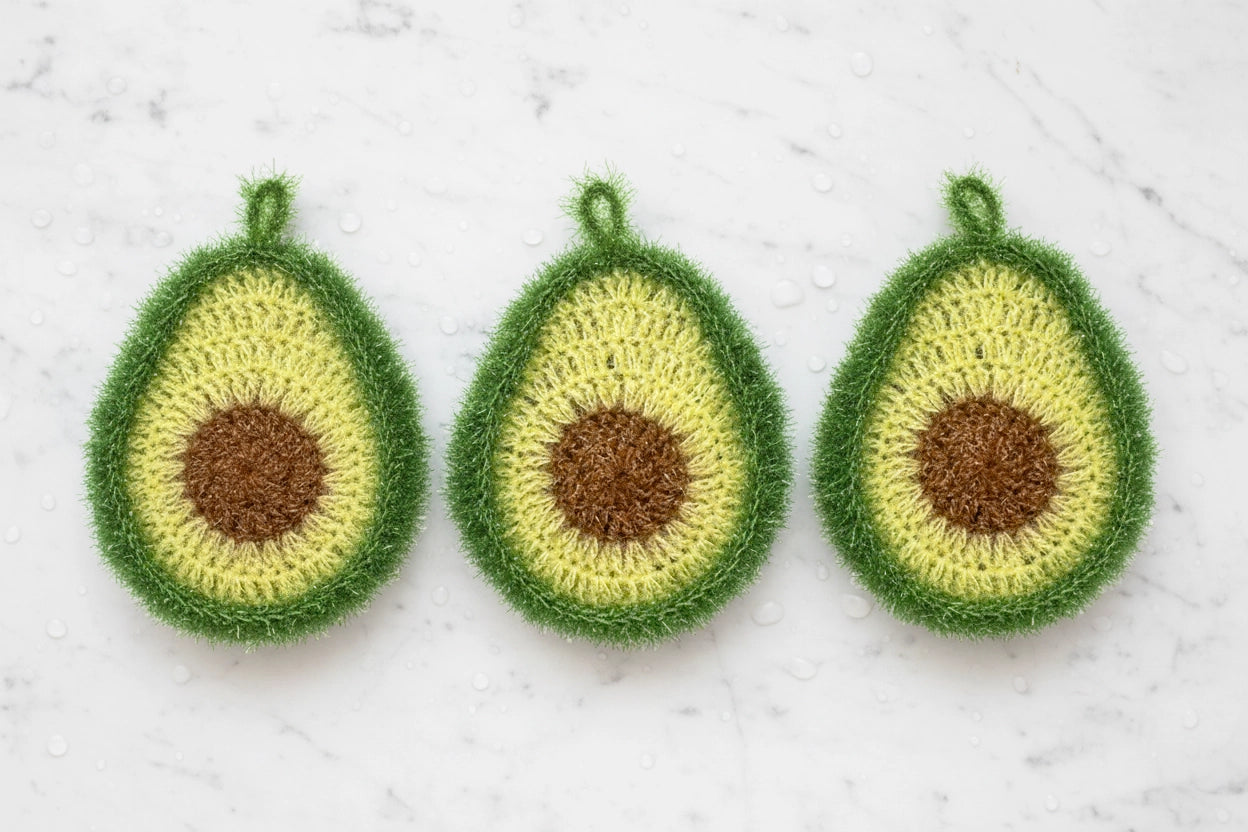 Three crocheted avocado-shaped dish scrubbers on a textured white marble background