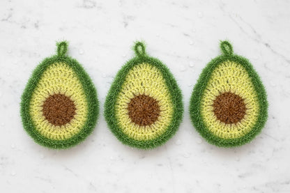 Three crocheted avocado-shaped dish scrubbers on a textured white marble background