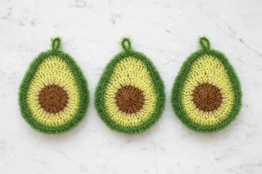 Three crocheted avocado-shaped dish scrubbers on a textured white marble background