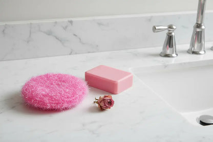 Pink scrubber, bar of soap, and dried rose on a marble countertop.