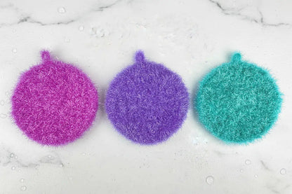 Three colorful Neon Collection scrubbies (magenta, purple, turquoise) on a marble surface.