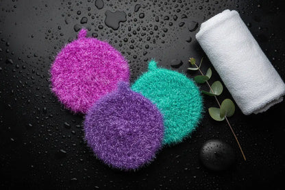Trio of colorful Neon Collection scrubbies (magenta, purple, turquoise) and a white towel on a black surface with water droplets.