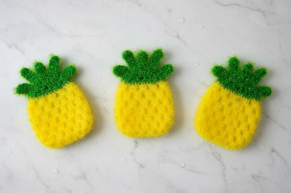 Three yellow pineapple-shaped crocheted dish scrubbers on a white marble surface