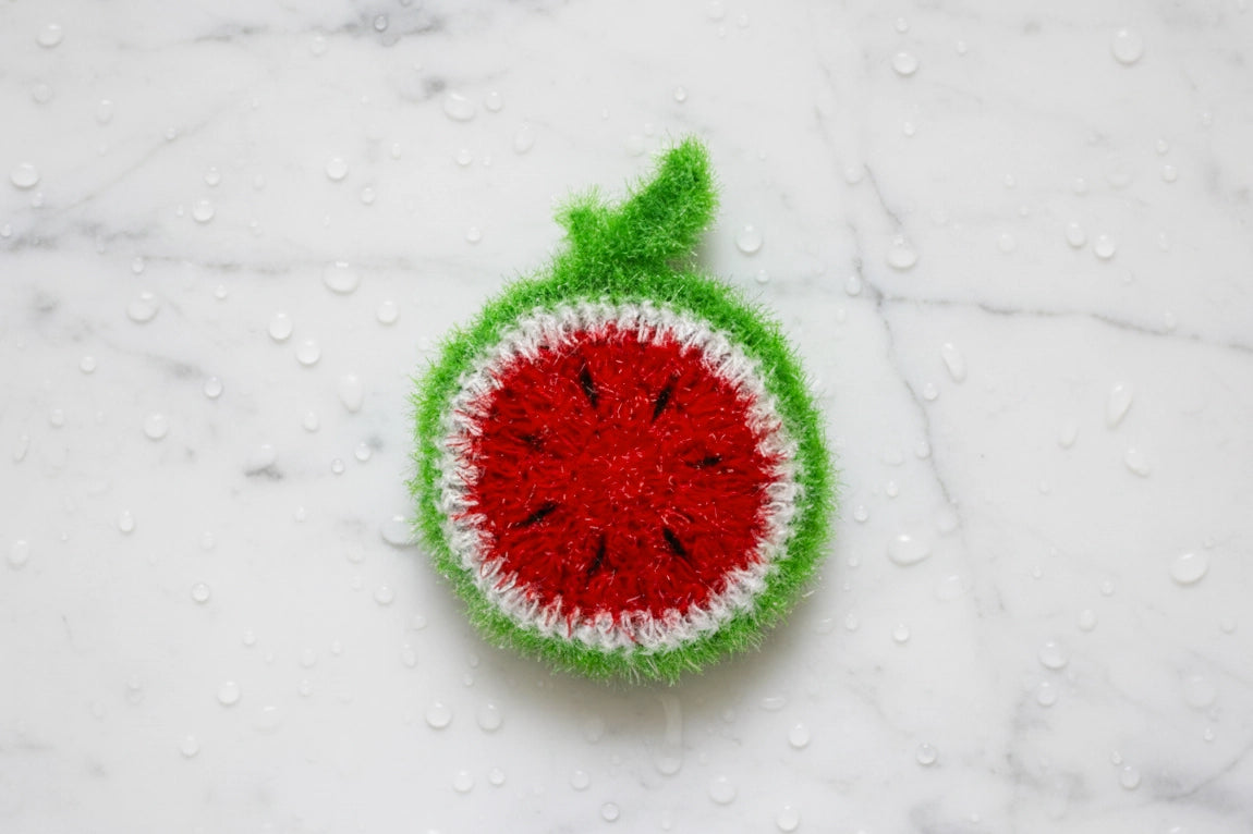 Top-down view of a crocheted watermelon dish scrubber on a white carrara marble surface