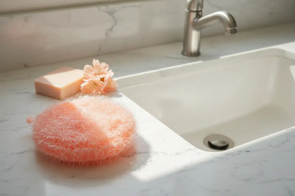 Peach Hello Scrubby on a marble countertop with soap and a faucet in the background.