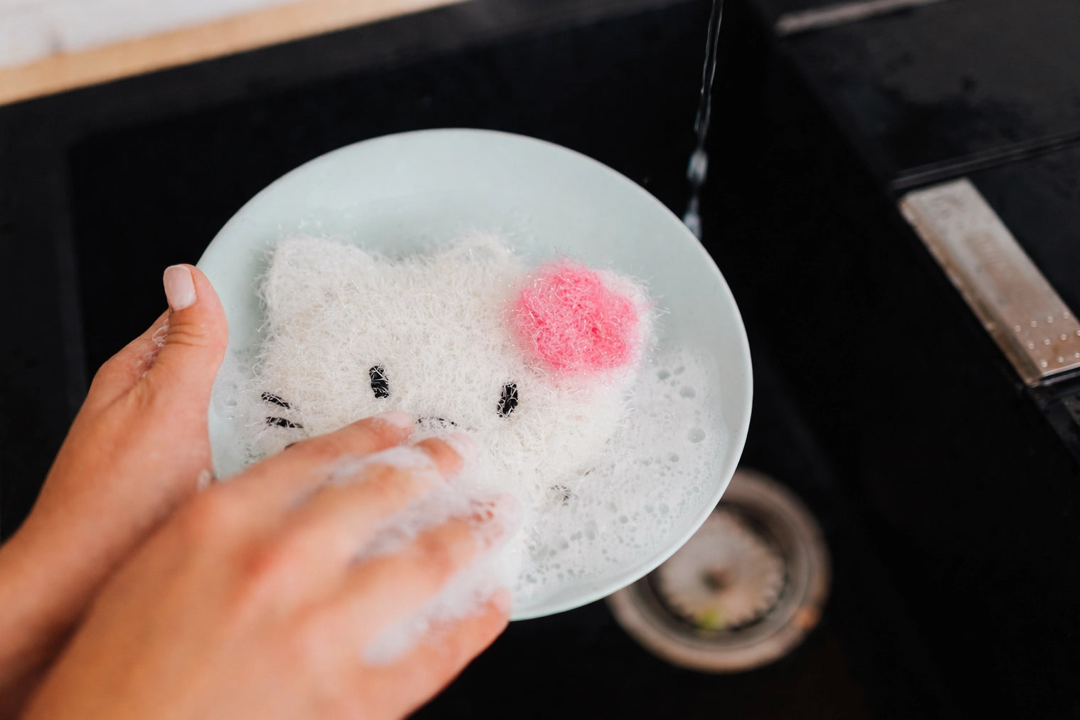 Person washing a white plate with a cat-shaped dish scrubber in a kitchen sink.