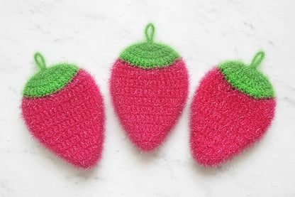 Three pink strawberry-shaped dish scrubbers with green tops on a white marble surface.