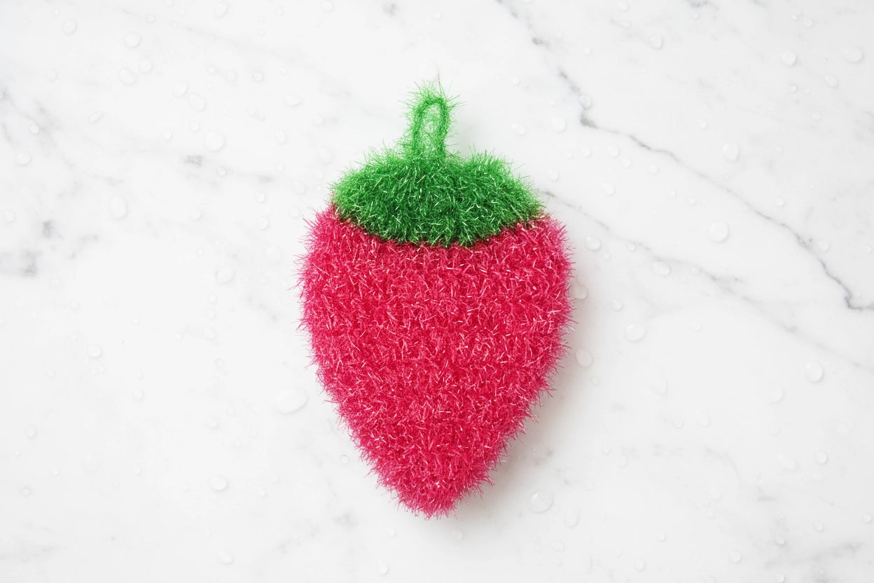 Pink strawberry-shaped dish scrubber with green top on a white marble background