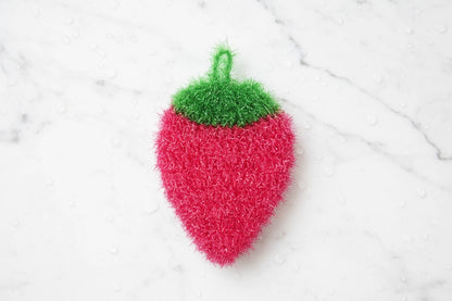 Pink strawberry-shaped dish scrubber with green top on a white marble background