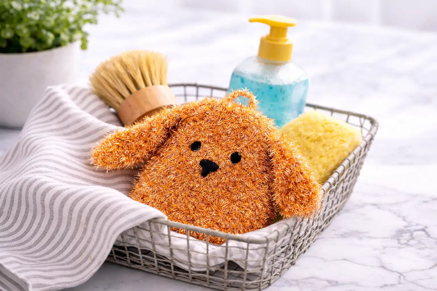 Basket with a brown dog-shaped dish scrubber, a brush, and a bottle of soap on a marble surface.