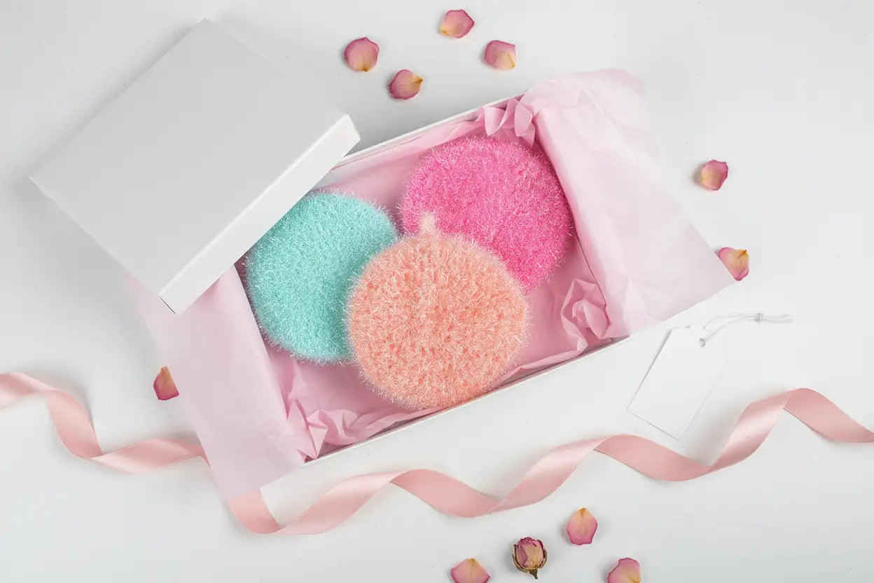 Three colorful scrubbies in a pink box with decorative ribbons and rose petals on a white background