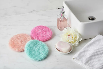 Three colorful scrubbies on a marble surface with a sink, soap, and towel in the background.