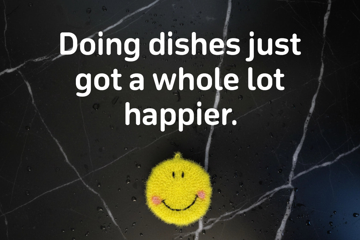 Smiley face crocheted dish scrubber on a textured black surface with text 'Doing dishes just got a whole lot happier.'