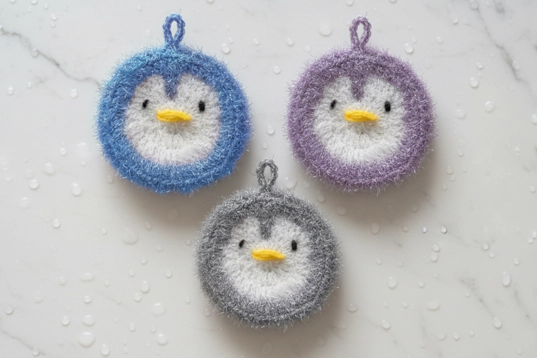 Three crocheted penguin dish scrubbers in blue, purple, and gray on a textured white background.