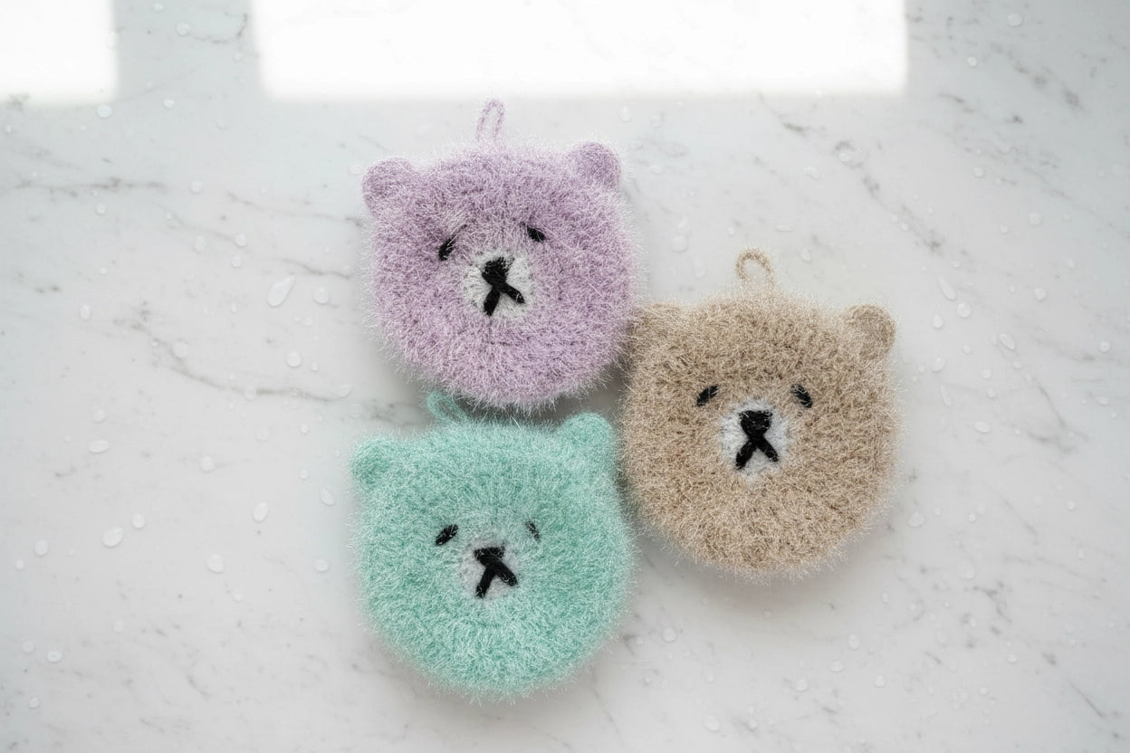 Three bear-shaped hand-crocheted dish scrubbers in purple, green, and light brown on a marble surface.