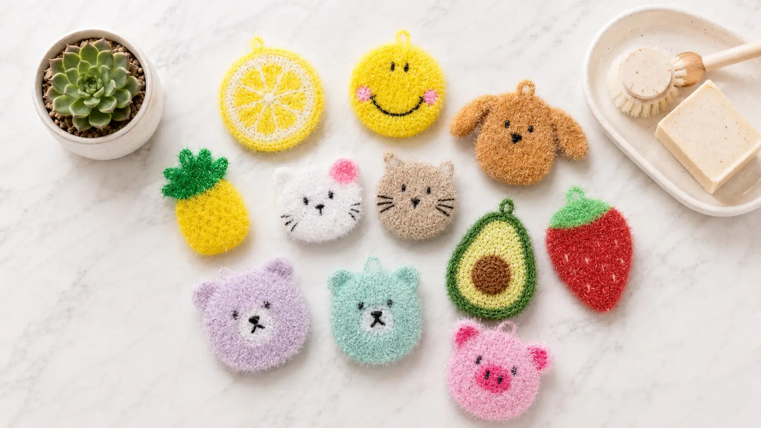 Colorful dish scrubbies shaped like animals and fruits on a white surface with a small plant and soap dish in the background.