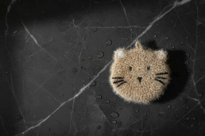Fuzzy cat-shaped dish scrubber on a dark, textured surface with water droplets.