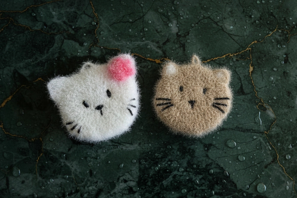 Two fluffy cat-shaped dish scrubbers on a dark textured surface