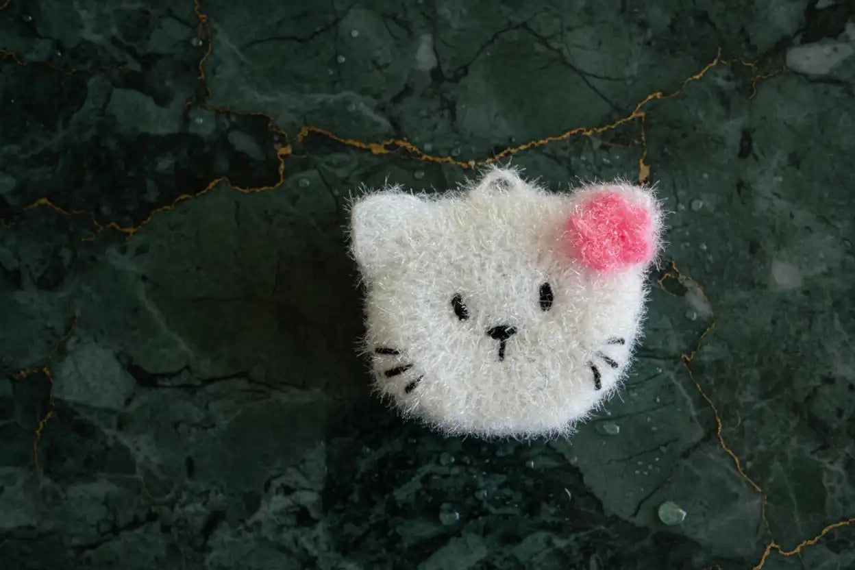 White cat-shaped dish scrubber with a pink flower on a dark textured surface