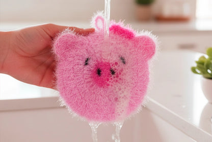 Hand holding a pink pig dish scrubber under running water, against a blurred kitchen background