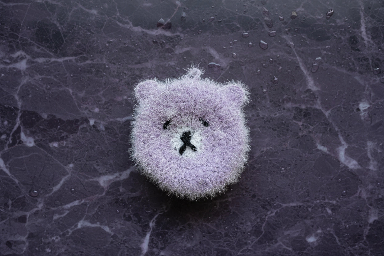 Fuzzy purple bear dish scrubber on a textured dark surface