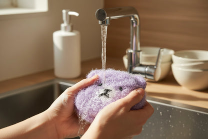 Purple bear dish scrubbers being rinsed under running water in a kitchen sink.