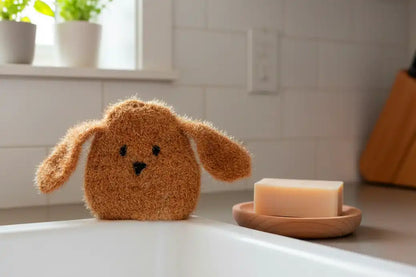 Brown dog-shaped dish scrubber on a kitchen sink countertop with a bar of soap on a wooden dish.