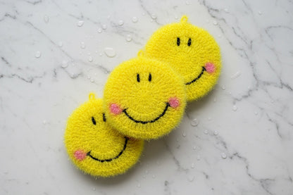 Three yellow smiley face dish scrubbers on a marble surface